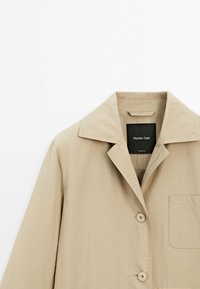 Beige jakke lavet af letvægtsstof. Har en bred krave, to knapper og en brystlomme. Glat tekstur med minimal detaljering.