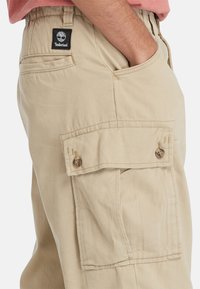 Beige cargo bukser med en lomme på låret, der har en knaplukning og et Timberland-logo mærke på taljen.