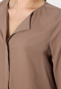 Blouse légère marron avec un col en V, texture lisse et éclat subtil, dotée d'une fermeture à boutons et d'un design à coupe décontractée.