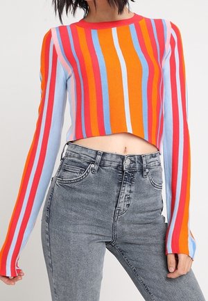Kobieta ubrana w krótki sweter w pionowe, wielobarwne paski z długimi rękawami i szare jeansy z wysokim stanem.