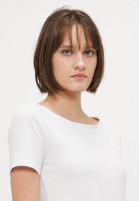 Hvit kortermet T-skjorte med bred hals, glatt tekstur og tettsittende design, båret av en person med brunt hår og blå øyne.