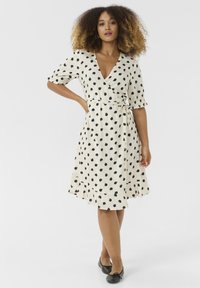 Knee-length wrap dress in cream with black polka dots, short puff sleeves, and flounce hem. Soft fabric with a tied waist detail.