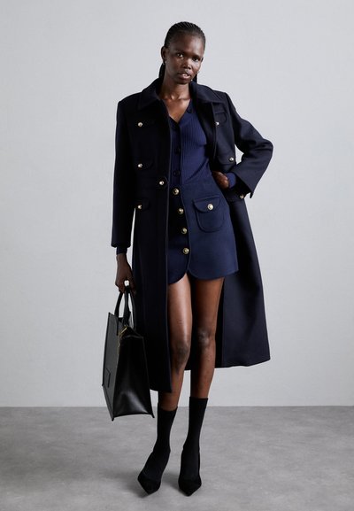 Navy wool coat with gold buttons, paired with a matching dress and black high heels. The model holds a black handbag. Textured knit top.