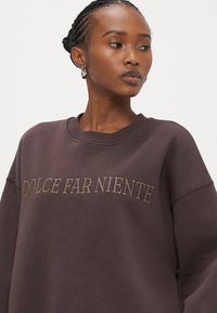 Tumši brūns džemperis ar izšūtu uzrakstu "DOLCE FAR NIENTE" gaišākā brūnā krāsā. Piedāvā ērtu, vaļēju piegriezumu un ribotu kakla izgriezumu. Gluda tekstūra.