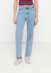 Hele sinised sirge lõikega teksapüksid, valmistatud denimist, millel on kõrge vöökoht, viis taskut ja pehme pleekinud efekt. Kandmas beeže jalanõusid.