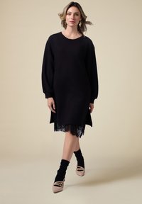 Schwarzes Sweatshirt-Kleid mit langen Ärmeln und Spitzenabschluss. Weicher Stoff mit geripptem Ausschnitt, kombiniert mit schwarzen Socken und spitzen beigen Schuhen.