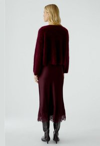 Bordeaux cropped fluffy trui over een aansluitende bordeaux jurk tot de vloer met kantafwerking aan de zoom, gecombineerd met zwarte kniehoge laarzen.