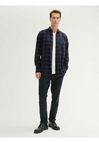 Granatowa flanelowa koszula w kratę z zapięciem na guziki, kieszenią na piersi, noszona na białym T-shircie, w zestawieniu z ciemnymi spodniami i czarnymi butami.