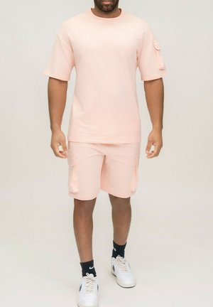 Homme portant une chemise à manches courtes rose clair et un short cargo assorti, avec des chaussettes noires Nike et des baskets blanches Nike.