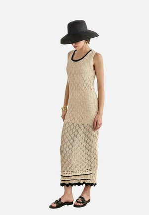 Mujer con un vestido maxi sin mangas de crochet beige con ribete negro, sombrero de ala ancha negro, sandalias negras y pulsera dorada, de pie y mirando ligeramente hacia la izquierda.