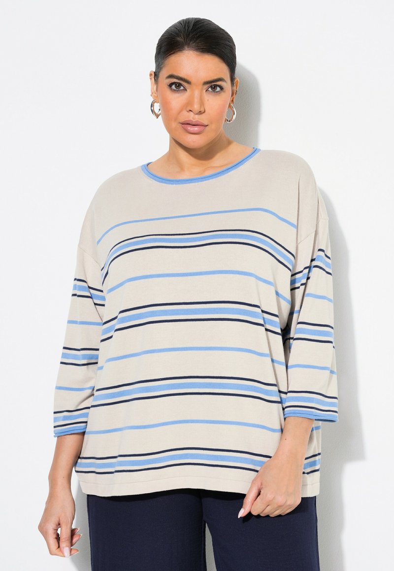 Nainen, joka pukeutuu vaaleanbeigeen neulepuseroon, jossa on vaaleansinisiä ja mustia vaakaraitoja, sekä tummansinihin housuihin, seisoo valkoista taustaa vasten.