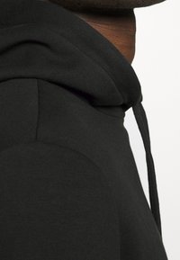 Sweat à capuche noir en tissu doux et texturé avec une capuche à cordon de serrage et une coupe ajustée, présentant des coutures courbes le long des côtés.