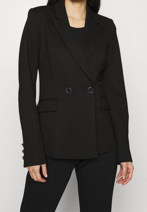 Mujer con un blazer negro entallado de doble botonadura con solapas de pico y bolsillos con solapa, combinado con pantalones negros sobre un fondo liso.