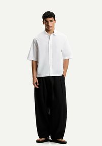 Camicia bianca a maniche corte, con fiocco rilassato, abbinata a pantaloni neri larghi e sandali neri. Tessuto liscio, design minimale.
