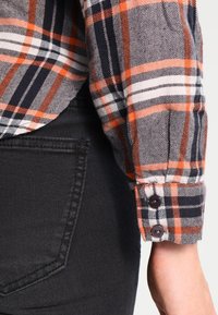 Manica di camicia a quadri in grigio, arancione e blu navy con polsino abbottonato; indossata con jeans neri. Presenta un tessuto strutturato e cuciture visibili.
