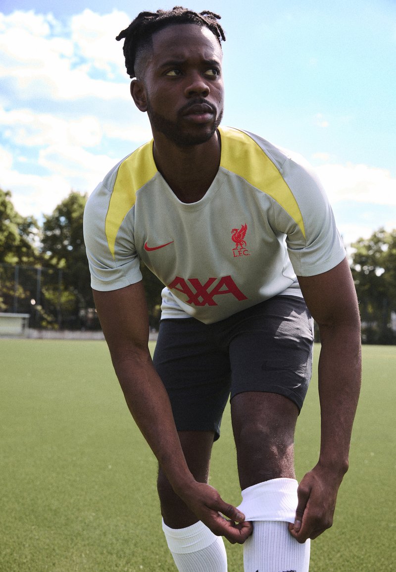 Gray Nike jersey with yellow accents and red Liverpool logo, paired with dark shorts and white socks, set on a green field.
