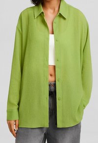 Camicia button-up verde, texturizzata, con colletto, maniche lunghe e vestibilità rilassata, indossata sopra un top corto bianco e abbinata a pantaloni grigio scuro.