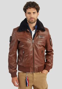 Veste aviateur en cuir marron avec un col en fausse fourrure noire, des poignets et un ourlet côtelés, des poches latérales et un patch brodé sur la poitrine.