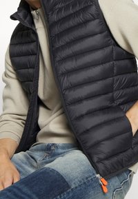 Mann trägt einen beigen Pullover, eine schwarze Steppweste mit orangefarbenen Reißverschlusszügen und patchworkblaue Jeans, sitzt mit den Händen nahe der Taille.