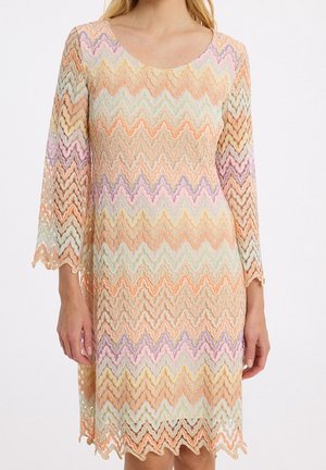 Femme portant une robe en dentelle à zigzag multicolore pastel avec des manches trois-quarts et un ourlet festonné, debout devant un fond blanc.