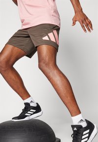 Pantalones cortos deportivos marrones con acentos triangulares rosas, zapatillas negras y calcetines blancos; individuo de pie sobre una pelota de estabilidad.