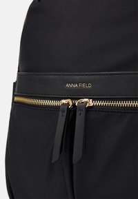 Sac à main en nylon noir avec une fermeture éclair dorée dotée d'un accent en cuir noir, un logo embossé et deux tirettes de fermeture éclair. Texture lisse sur toute la surface.