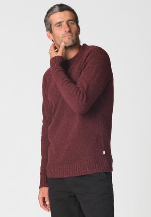 Homme portant un pull en maille bordeaux et un pantalon noir, touchant son cou d'une main sur un fond clair uni.