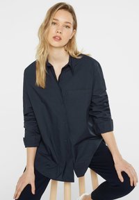 Navy blue button-up shirt with a collar, single chest pocket, rolled sleeves, and smooth texture, paired with dark trousers.
