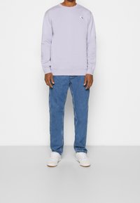 Felpa lilla con collo a giro, polsini a costine e un piccolo logo ricamato. Abbinata a jeans in denim blu e sneaker bianche.