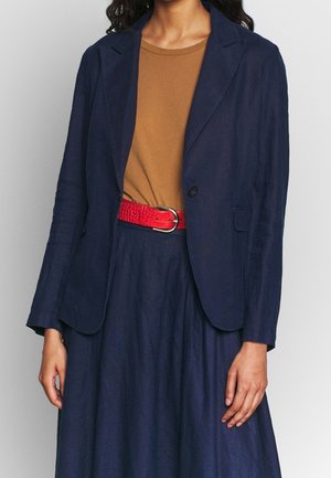 Kvinde iført brun skjorte, navy blazer, navy nederdel og rødt bælte med sølvspænde, vist fra hals til midt på låret.
