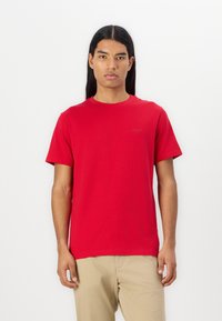 Camiseta de algodón roja con un cuello redondo clásico, mangas cortas y un logotipo sutil en un tono más oscuro en el pecho. Textura suave.