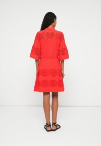 Vestuário vermelho com um silhueta larga, mangas curtas e padrões recortados intrincados ao longo das costas e do fundo. Textura de tecido suave.