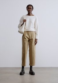 Fehér, hosszú ujjú pulóver, bézs, magas derekú nadrág és fekete platform bokacsizma. Egyik kézben egyszerű fehér táska. Minimalista háttér.