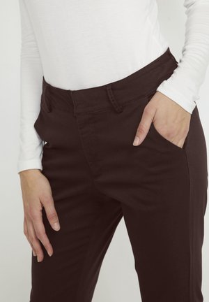 Persoon draagt donkerbruine strakke broek met hand in zak en een wit shirt met lange mouwen tegen een effen lichte achtergrond.