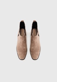Botas de tobillo de ante marrón con una textura suave, punta afilada, paneles laterales elásticos y suelas negras planas, con detalles de costura sutiles.