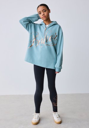 Junge Person trägt einen türkisfarbenen, übergroßen Hoodie mit dem Schriftzug "Baker", schwarze Leggings mit dem gleichen Schriftzug und weiße Sneaker und steht vor einer schlichten Wand.