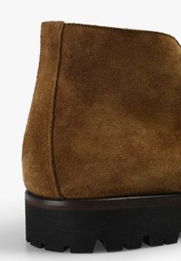 Brune ruskindsankelstøvle med en tekstureret overflade, en stablet sort gummisål og en glat træaccent på hælen.