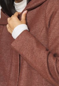 Abrigo de color óxido con un tejido texturizado, que cuenta con capucha y un ajuste ceñido. Mano agarrando la abertura delantera.
