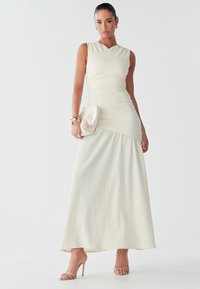 Une femme se tient avec confiance dans une robe ajustée couleur crème, tenant une petite pochette beige et portant des sandales à lanières, avec des cheveux lisses et des bijoux.