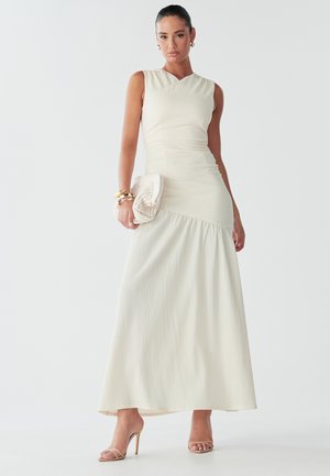 Kobieta stoi pewnie w dopasowanej, kremowej sukience, trzymając małą beżową torebkę i nosząc sandały na obcasie, z gładkimi włosami i biżuterią.