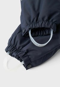 Jūras zila virsjaka ar elastīgām piedurknēm, kurai ir caurspīdīgas plastmasas gredzeni un melni pogas ar spiedienu regulēšanai. Gluda auduma tekstūra.