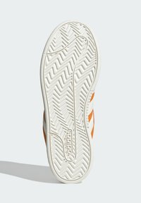 Suela de zapatilla blanca con patrón de zigzag, con franjas naranjas en los costados y la marca "adidas". Goma texturizada para mayor tracción.