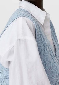 Zbliżenie na osobę noszącą białą koszulę z kołnierzykiem pod jasnoniebieskim kamizelką bez rękawów o splocie warkoczowym.