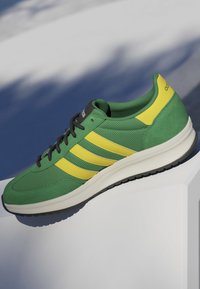 Green and yellow Adidas sneaker with black laces, white sole, and textured fabric, displayed on a white angled surface with shadowed background.