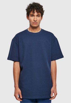 Tamno plavi oversized majica od teksturirane tkanine, s okruglim izrezom i kratkim rukavima. Ravni rub s minimalnim dizajnerskim detaljima.