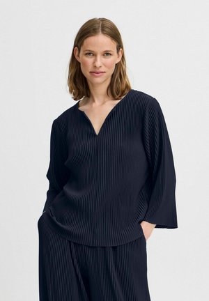 Navy blue pleated blouse with a V-neck and long sleeves, featuring a smooth texture and relaxed fit. No visible hardware or patterns.