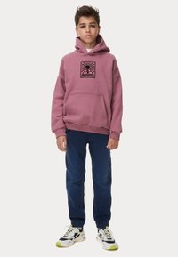 Sudadera rosa con un diseño gráfico, bolsillo frontal y puños acanalados. Combinada con vaqueros azul oscuro y zapatillas negras/blancas con acentos verdes.