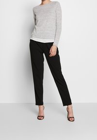 Suéter de manga larga gris claro con escote redondeado, superpuesto a una blusa blanca, combinado con pantalones negros ajustados y tacones negros con tiras.