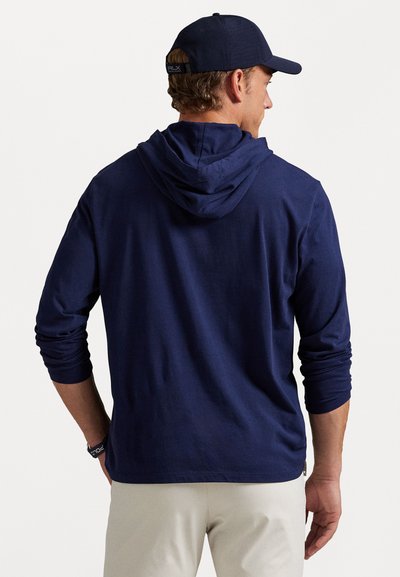 Man in een donkerblauwe hoodie en bijpassende pet, van achteren gezien, met beige broek tegen een effen witte achtergrond.