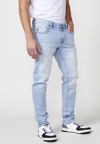 Jeans de denim azul claro con un corte slim, con cinco bolsillos, un ligero jacquard y una textura deslavada, combinados con zapatillas en blanco y negro.